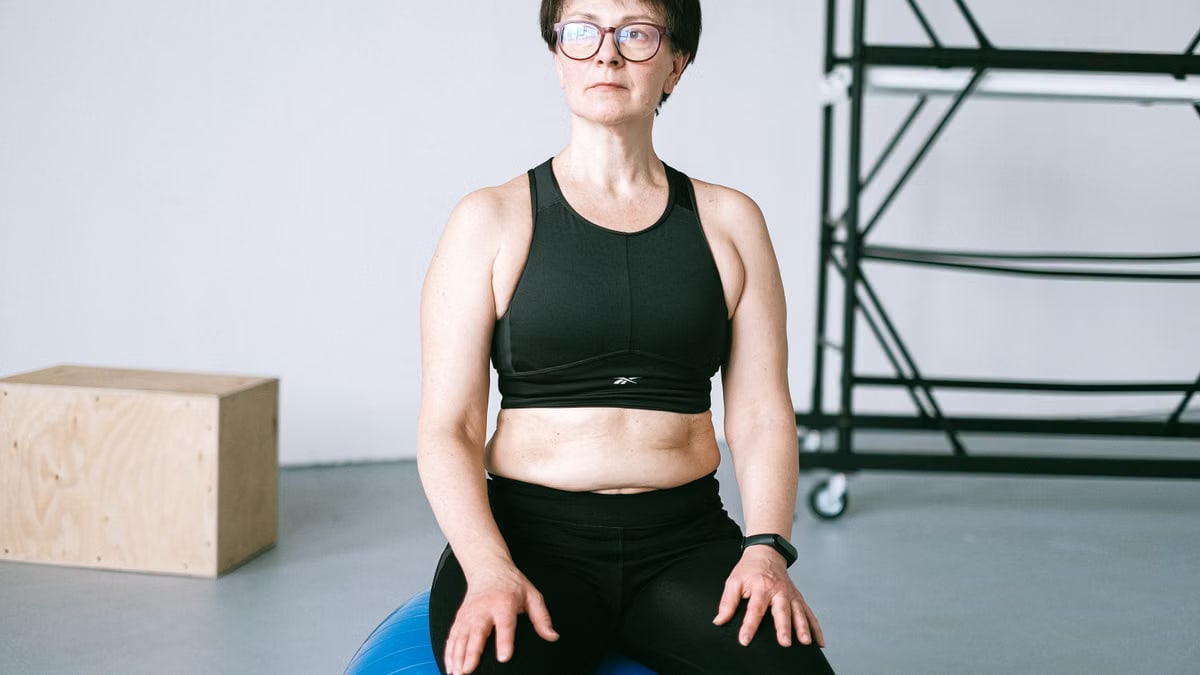 Middle-aged woman with eyeglasses exercising on a blue stability ball indoors.